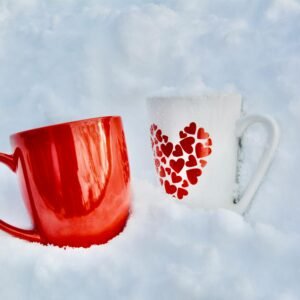 Two mugs in snow: a red mug and a white one with heart designs, evoking warmth and winter themes.